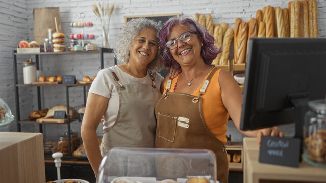 Two smiling middle-aged women bakers standing together in their bakery shop filled with various breads and pastries, creating a warm and welcoming atmosphere.