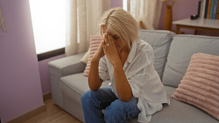 Woman sitting on couch in a living room with hands covering face, appearing stressed, wearing white blouse and jeans, in a cozy home environment with pink cushions and soft lighting