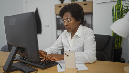 Middle-aged african american woman with curly hair working on a computer in an office indoor setting, focused and professional