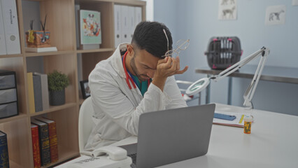 A handsome hispanic man with a beard, wearing a white lab coat, looking stressed in a veterinary clinic, sitting at a desk with a laptop, prescription bottle, and medical tools surrounding him.