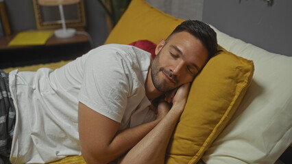 A handsome young hispanic man with a beard sleeps peacefully on a bed in a warmly lit bedroom, wearing a white shirt, surrounded by cozy yellow pillows and soft blankets.