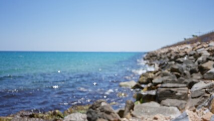 Blurred, outdoor scenery with a rocky shore and clear blue ocean under a bright sky showing defocused and bokeh effects in the background