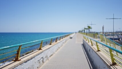 Obraz premium Blurred seaside promenade with defocused railings, pathway, and bokeh ocean view under clear blue sky