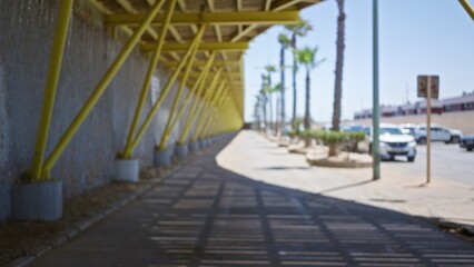 Fototapeta premium Blurred view of an outdoor pathway with yellow structural beams, palm trees, and parked cars.