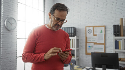 Mature hispanic man wearing red sweater using smartphone in modern office with white walls, clock, and documents displayed, creating a focused and professional indoor setting