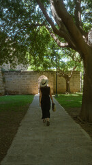 Woman walking under trees on a stone path in a sunny park in palma, mallorca, spain, wearing a hat and holding a handbag near old stone walls and an arched gateway