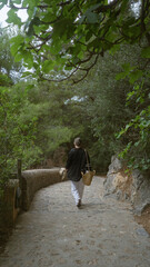 Young woman strolling along a scenic path surrounded by lush greenery in mallorca, spain, holding a hat and a woven bag.
