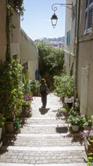 Woman exploring narrow european alley surrounded by lush plants on sunny day in charming cityscape with old lanterns casting shadows and historic stone path.