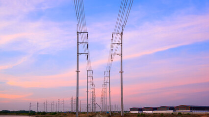 Row of electric poles and cable lines with industrial factory buildings and construction site in industry settlement area against evening sky background