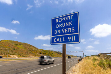 A blue traffic sign stating Report Drunk Drivers call 911 with cars driving by