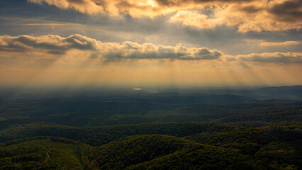 Golden sunlight filters through clouds, casting dramatic rays over a vast forest landscape at sunset, creating a breathtaking view