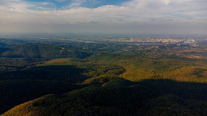 Fototapeta premium Aerial view of budapest city emerging from behind rolling green hills, showcasing urban development amidst natural landscape under a partly cloudy sky