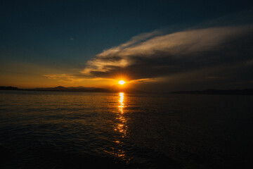 Golden hues of a setting sun paint the sky and reflect on the tranquil ocean, creating a serene scene with clouds and distant mountains