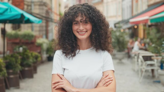 Charming Caucasian girl standing in middle of street while looking at camera. Crossing her hands and smiling. Wearing white shirt. Long curly hair. Many restaurants opened around. Suny weather.