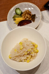 A bowl of steamed rice mixed with sweet corn is served alongside a small plate of colorful Japanese pickles, including cucumber, radish, and beans, creating a balanced and traditional meal.
