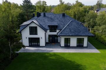 Obraz premium Modern house with dark gray slate roof. Large white house with black framed windows. Patio area with gray stone paving. Green lawn surrounds house. Trees, green plants visible in background.