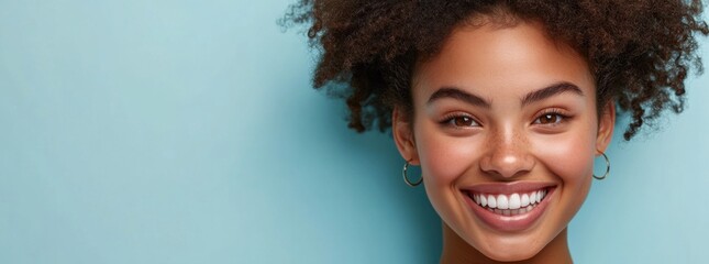 Young woman with curly hair smiles brightly, displaying her healthy teeth against a light blue background, emphasizing dental care and confidence