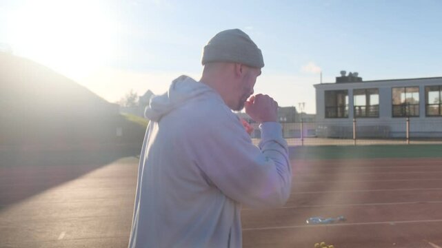 Man in gray hoodie and beanie practices boxing outdoors on a running track in bright sunlight