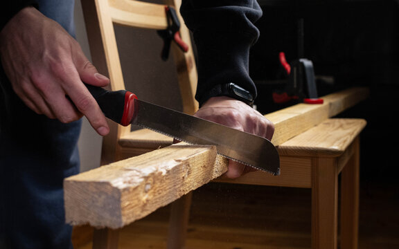 Close-up of a carpenter's hands using a hand saw to cut a wooden plank, showcasing woodworking skills and craftsmanship in a workshop setting.
