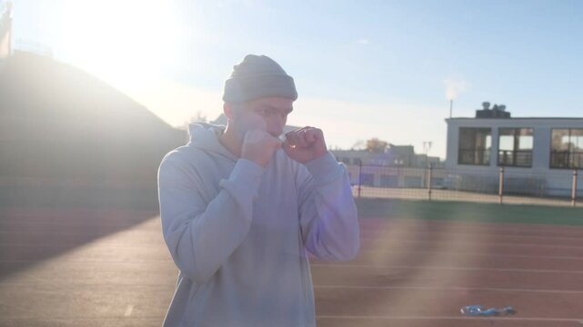 Man in gray hoodie and beanie practices boxing outdoors on a running track in bright sunlight
