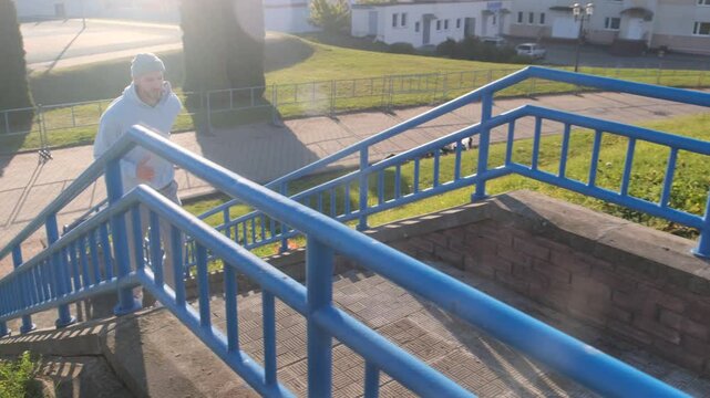 An athlete in a grey hoodie and trousers runs up a blue staircase doing outdoor exercises on a sunny day
