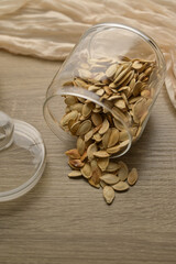 Scattered pumpkin seeds, in transparent glass jar on wooden table, beige cloth on the background