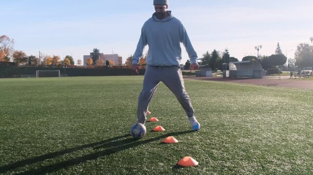 Football player practices dribbling. Man in blue hoodie, grey pants, controls the ball between cones. Sunny day, training on the field.