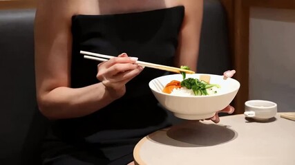 Elegant Dining:  A woman in a black dress enjoys a bowl of Japanese cuisine with chopsticks in a modern cafe setting. Her elegant demeanor adds a touch of sophistication to the scene. 