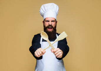 Angry bearded male chef, cook or baker in uniform with wooden spoon and spatula in hand. Cooking...