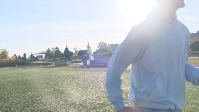 Man jogging on a sports field. He wears a gray hoodie, sweatpants and beanie. It is a sunny day