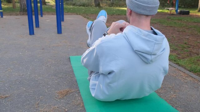 Man doing crunches on mat outdoor in park. Grey hat, blue sweatshirt, grey pants, blue sneakers. Sport, workout, exercise, fit