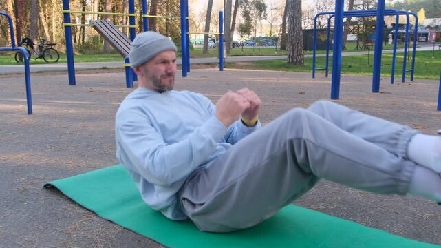 Man doing sit ups on mat outside in park, grey hat, blue sweatshirt, grey pants. Sport, workout, active, healthy