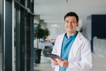 Smiling young male doctor holding a tablet and standing in a clinic hallway, looking at the camera