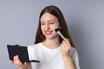 Smiling teenage girl applying blusher with makeup brush on grey background