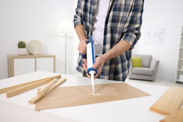 Man with caulking gun glueing plywood at white table indoors, closeup
