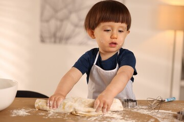Cute little boy kneading dough at wooden table indoors