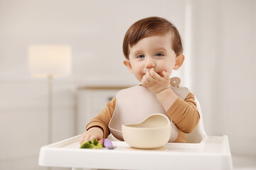 Cute little baby eating healthy food from bowl in high chair at home