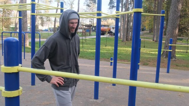 Man with hoodie walking through outdoor gym equipment. Trees, park in background. Fitness area. Autumn day