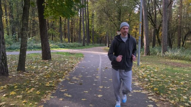 Man jogging on autumn park path. He is wearing grey hoodie, pants and hat