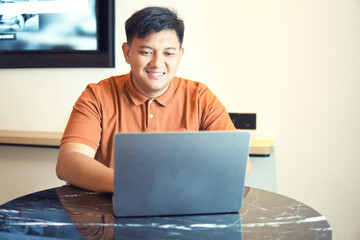 A man in an orange shirt is sitting at a table, working on a laptop. A young man smiling while working on a laptop in a casual home or office setting