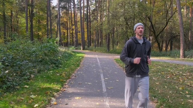 A man jogging in an autumn park. The path is paved, bordered by trees and fallen leaves. He is dressed in casual sportswear