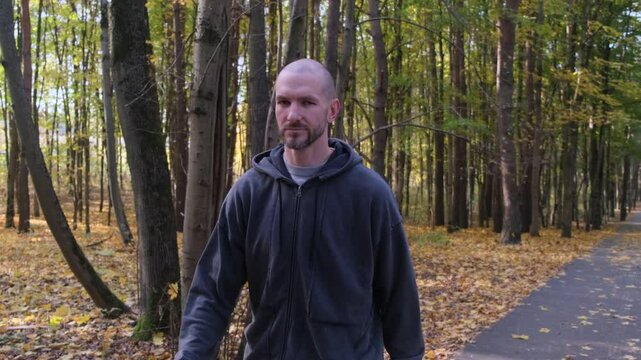 A pensive bald man in a grey hoodie walks through an autumn forest