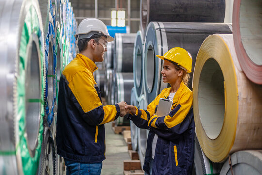 Two young engineers or mechanical workers in safety suit discuss and consult each other for their work routine next to the rolls of metal sheets in a factory. Partnership among two workers.