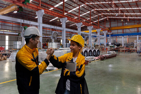 Two young engineers or mechanical workers in safety suit celebrate their agreement with high five next to the rolls of metal sheets in a factory. Partnership among two workers. Cooperation in factory