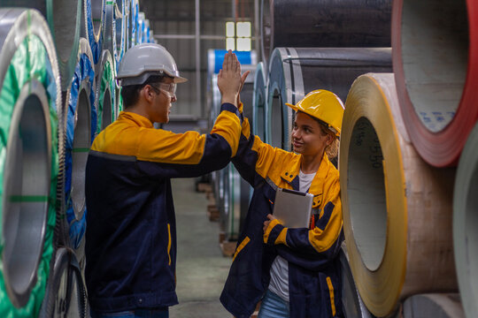 Two young engineers or mechanical workers in safety suit celebrate their agreement with high five next to the rolls of metal sheets in a factory. Partnership among two workers. Cooperation in factory