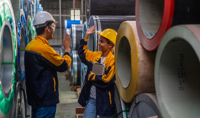 Two young engineers or mechanical workers in safety suit celebrate their agreement with high five next to the rolls of metal sheets in a factory. Partnership among two workers. Cooperation in factory