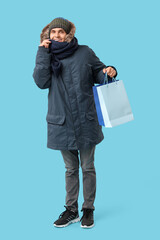 Young man in stylish puffer jacket and scarf with shopping bags on blue background
