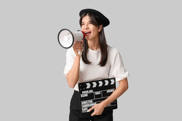 Young happy female film director with movie clapper and megaphone screaming on grey background