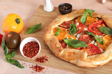 Wooden board of tasty tomato galette with basil and spices on beige background