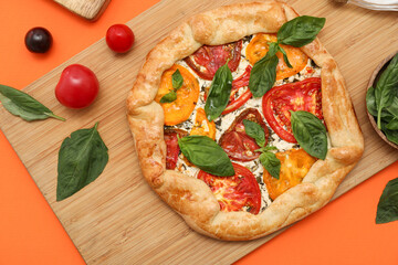 Wooden board of tasty tomato galette with basil on orange background